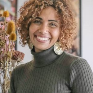 Image of a woman with curly hair and a green jumper standing by some dried flowers.
