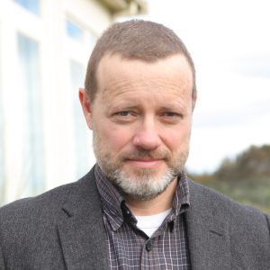 Image of a man with short hair and beard and grey jacket looking at camera.