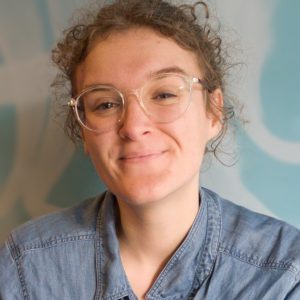 Image of a woman with curly hair and glasses and a denim shirt smiling at the camera.
