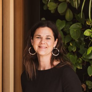 Image of a woman with dark hair and hoop earrings standing against a wooden wall.