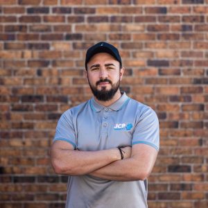 Image of a man in a cap and grey shirt standing with arms folded in front of a brick wall.