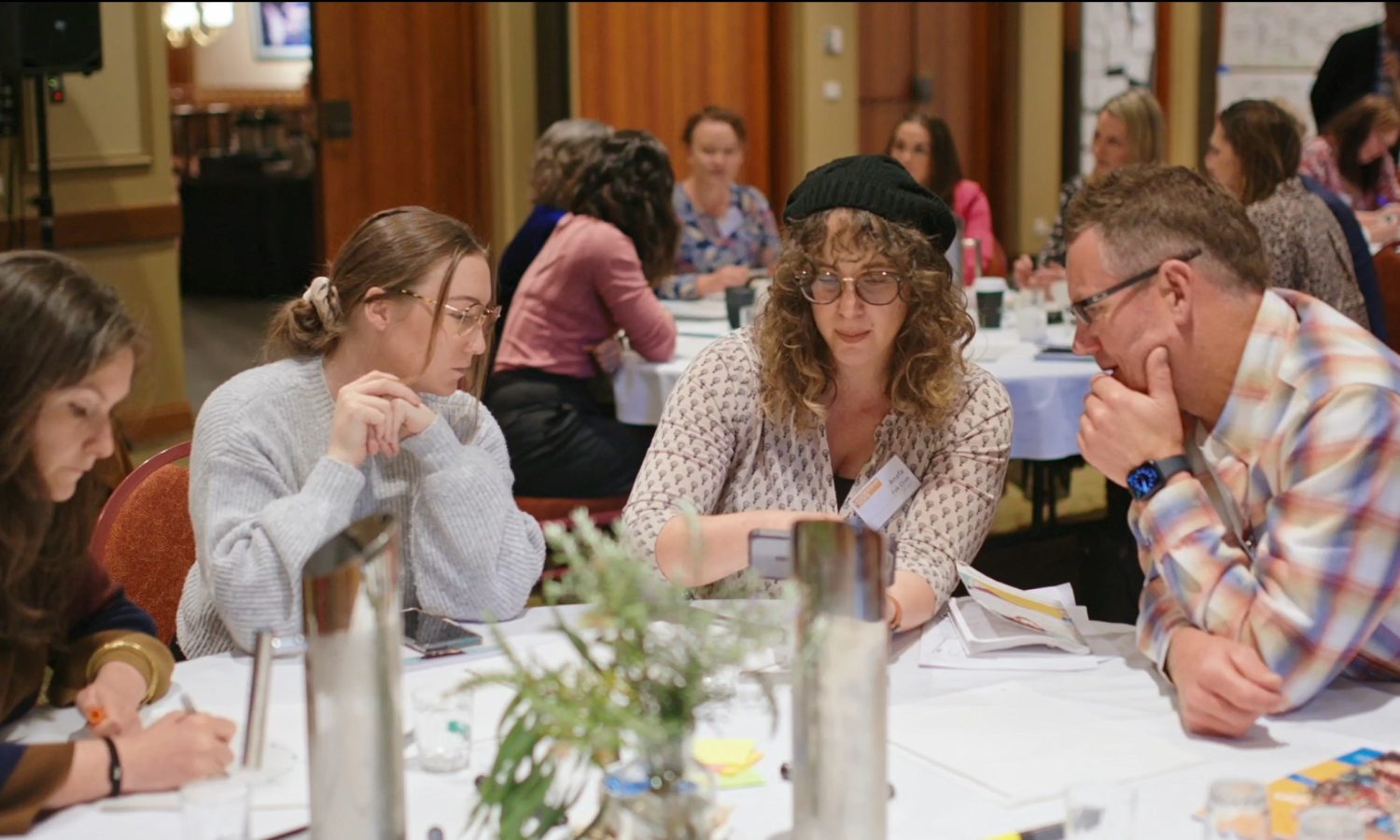 image of participants sitting around a table looking at something together. other participants at another table behind them.