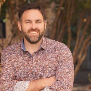Image of a man with his arms crossed in a colourful shirt with short dark hair and a beard, trees in the background.