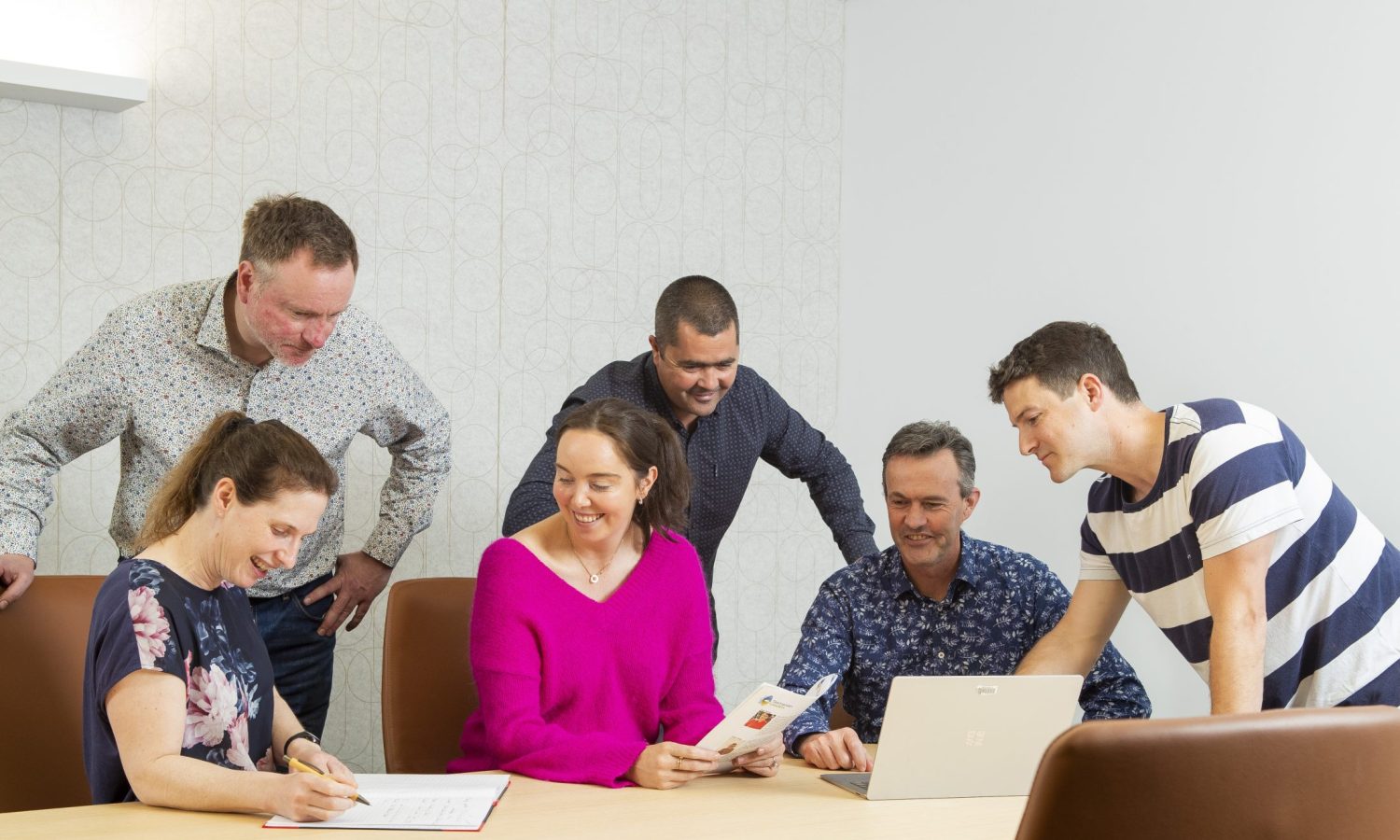 Image of a group of 6 working together. Some are using a lap top, one is writing and one is reading a document. They are all smiling and look engaged.