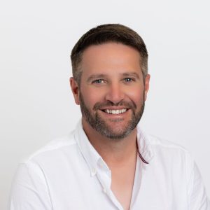 Image of a man with short dark hair and a cropped beard wearing a white shirt. Background it white.