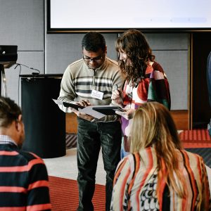 A photo of a man and a woman standing looking at documents together . They stand amongst others working who are both sitting and standing, in front of a stage with lectern and microphone.