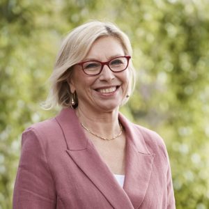 Image of a woman with short blonde hair and red framed glasses smiling at the camera. She wears a pink jacket and there is greenery in the background.