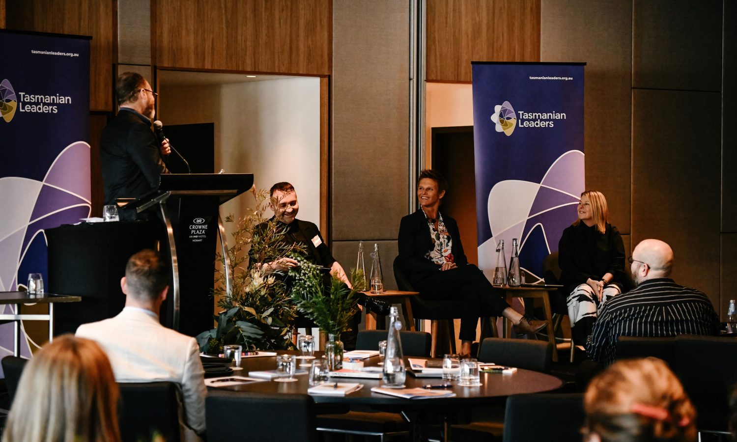 Image of 3 seated panel members interacting with facilitator. Members of the audience in the foreground.