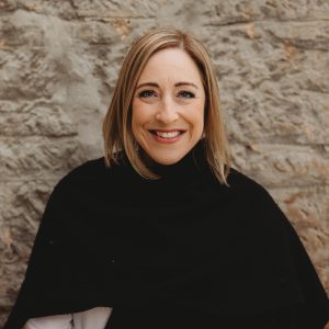Headshot of a woman standing against an old stone wall wearing a black top. She smiles directly at the camera.