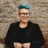 Head shot of Katy Cooper. Katy has a black jacket and short spikey green hair and glasses