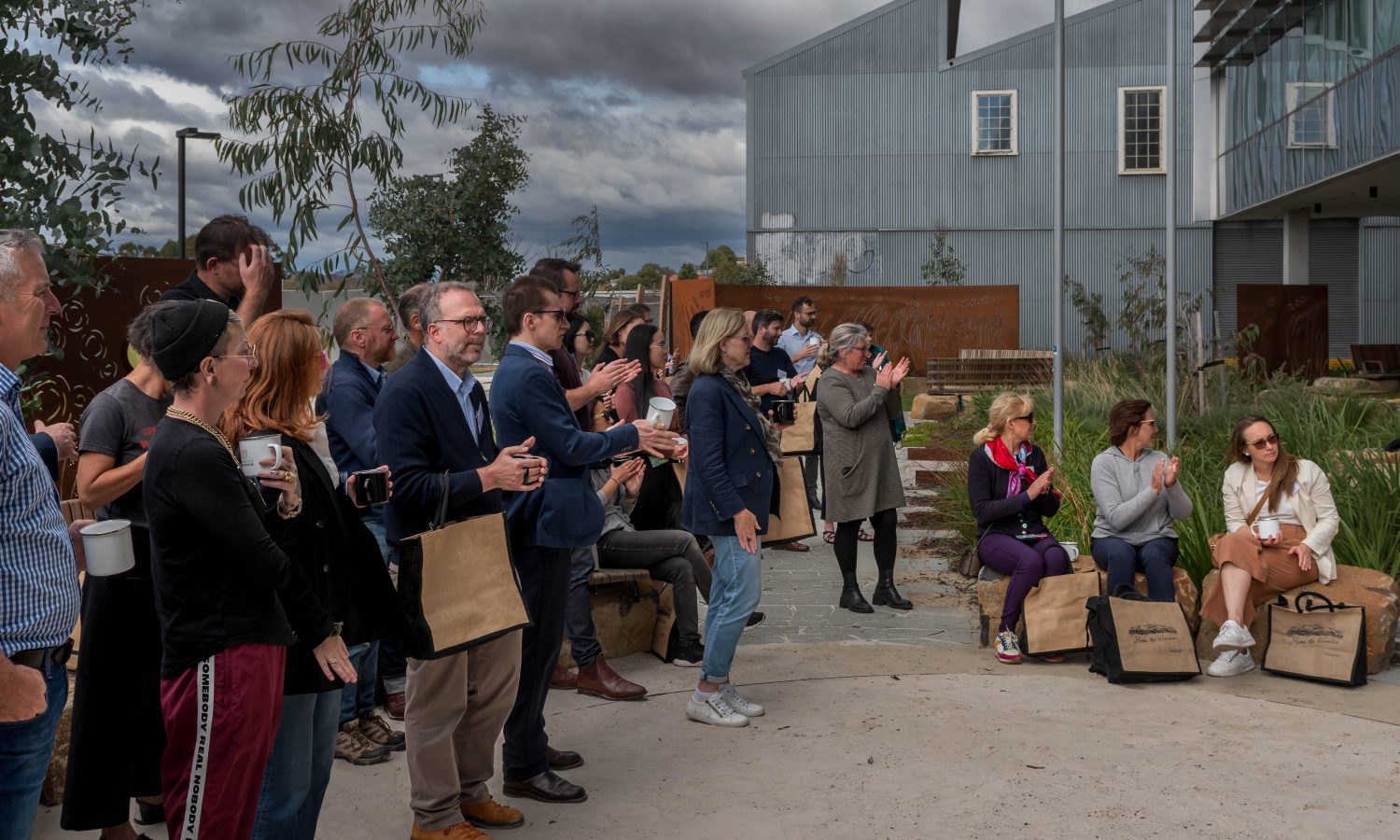 Image of a group of people standing and sitting in a garden, clapping. The bag ground is a larch steel structure and a moody cloudy sky.