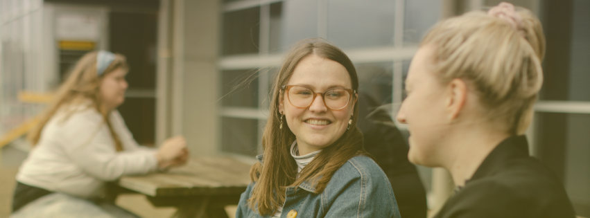 Image of two women sitting outside talking. There is another woman in the background sitting at a table alone.