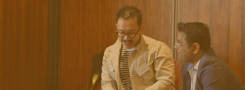 Image of two men sitting against a wooden wall, discussing ideas