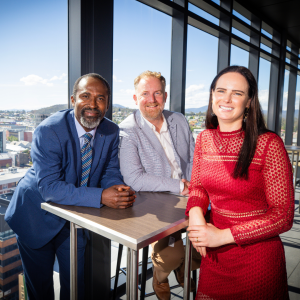 Photo of three TLP participants from 2023. A woman with long black hair and a long sleeved red dress, a fair haired man with a beard in a grey suit jacket and a dark haired man in a blue suit leaning on a small table together. View behind them overlooks the city of Hobart.