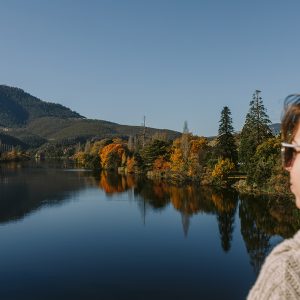 Image of a woman looking over a calm body of water surrounded by trees in Autumn colours.