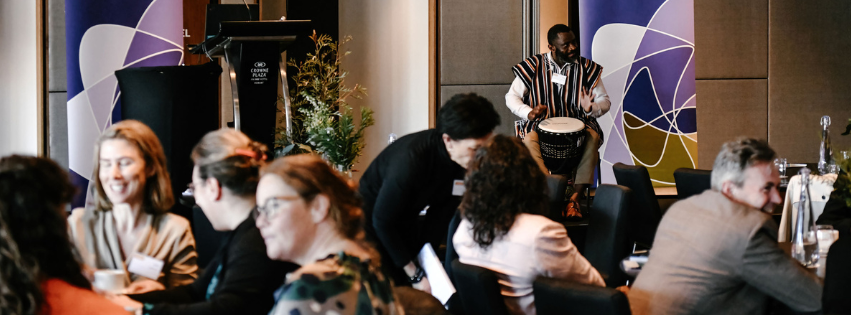Image of someone playing hand drums on stage while audience talk, smile and look engaged with each other.