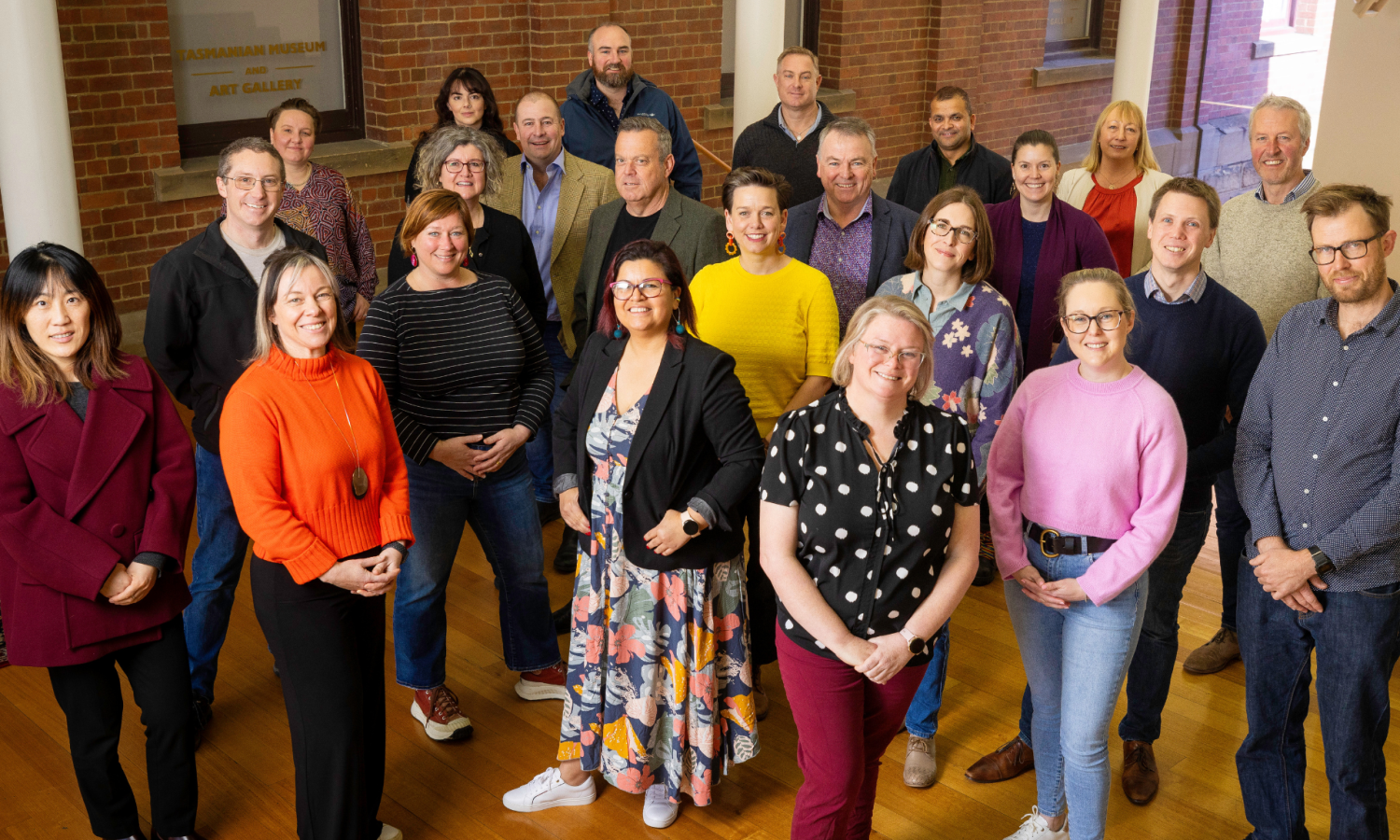 A landscape group photo of 23 of the 2024 Tasmanian Leaders program participants. All wearing brightly coloured professional wear, all smiling and looking directly at the camera. Brick wall background.
