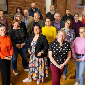 A landscape group photo of 23 of the 2024 Tasmanian Leaders program participants. All wearing brightly coloured professional wear, all smiling and looking directly at the camera. Brick wall background.