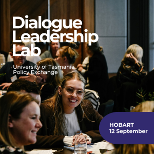 Photo is of people sitting at round tables at an event, most are talking amongst themselves and girl in centre of photo is smiling happily and looks engaged in conversation. She has long blonde hair, glasses, white shirt and brown jacket. the others in the photo are blurry.