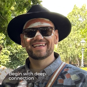 A photo of a man in a hat and sunglasses with First Nations face paint on his forehead and nose. He is smiling at the camera and standing in front of green trees.