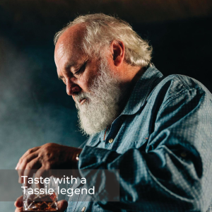 Image of a man with white hair and white bushy beard wearing a blue checked shirt holding and looking down at a glass of whisky.