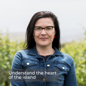 A portrait picture of a woman with a denim button up shirt, long black hair and glasses, small pink earrings and backdrop is blurred bushes and a grey sky.