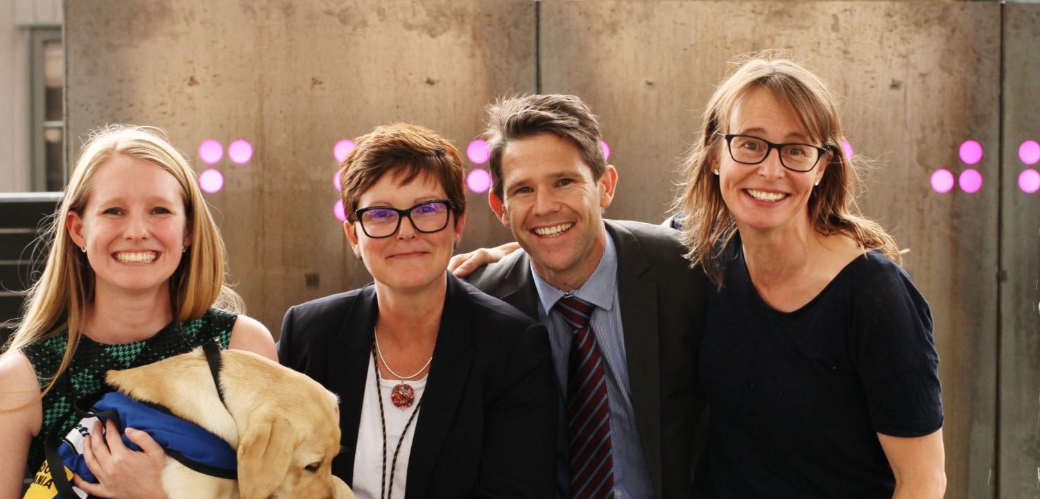 A landscape photo of 5 people. L-R woman with long grey cardigan with large silver necklace and shoulder length curly black hair, Woman with long blonde hair holding a guide dog puppy, A woman with short dark hair and glasses, a man in a suit leaning in with short spikey brown hair and a taller woman at the end with a black top, long hair and glasses. All are smiling directly at the camera.