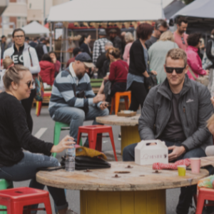 A landscape shot of the Launceston Harvest Market. It is busy with lots of people around, the centre of the photo is a family sitting at a wooden table with green and red stools relaxing and chatting.
