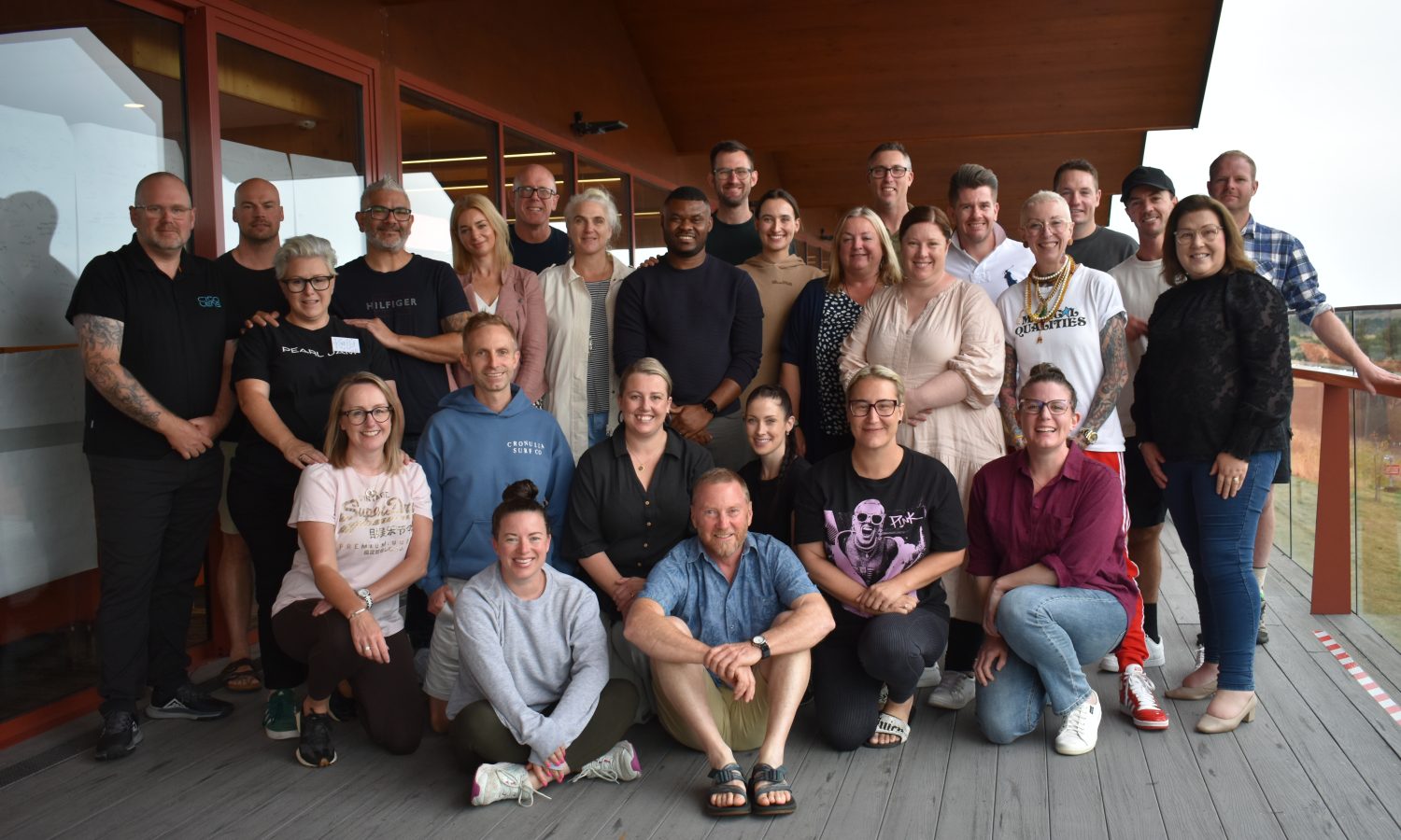 A group photo of 2025 Tasmanian Leaders Participants standing on a deck under a roof, in casual clothes all looking at the camera smiling. Some are sitting cross legged, some are kneeling and others standing at the back.