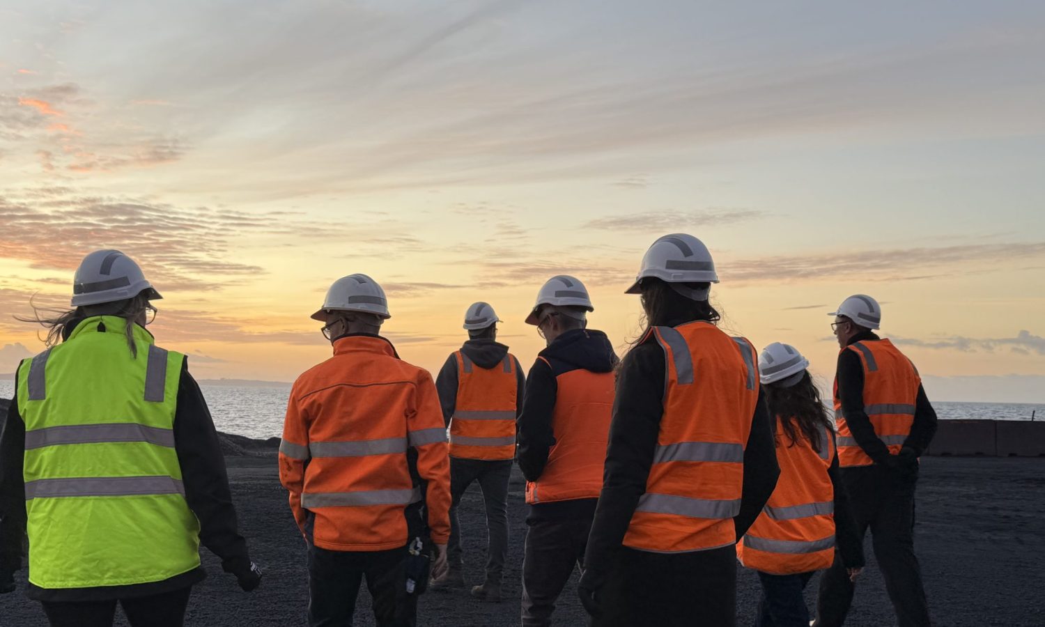 Tasmanian Leaders participants at sunset walking on gravel in high vis vests and hard hats. all have their back to the camera. Sky is orange with ocean in the background.
