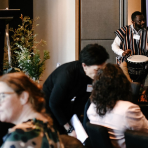 A landscape photo of a man playing hand drums on stage in front of members of the Symposium audience. There are Tasmanian Leaders banners and a lectern in the background.