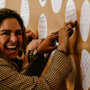 A landscape image of a man and a woman sticking round stickers onto a brown paper covered wall. The woman is smiling excitedly at the camera and the man wearing glasses is focused on the wall.