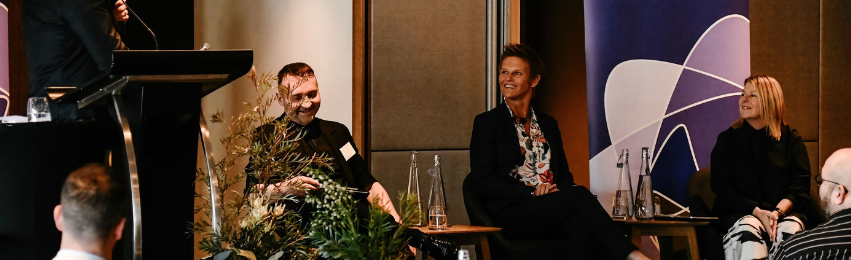 Panel photo of three panellists a man with short dark hair and a dark jumper, a woman with short spikey hair and a floral shirt and blonde woman wearing black and white. All are seated on a stage and smiling.