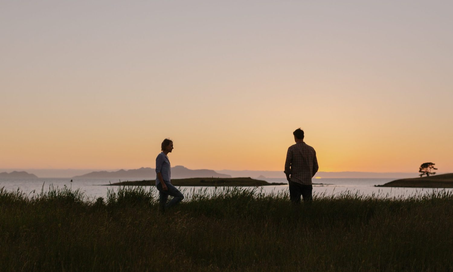 A photo taken as the sun sets on a hill overlooking a bay with islands. Two silhouettes one of a man with hands in his pockets looking at the view and a woman walking through long grass to meet him. They sky is a light orange at dusk.