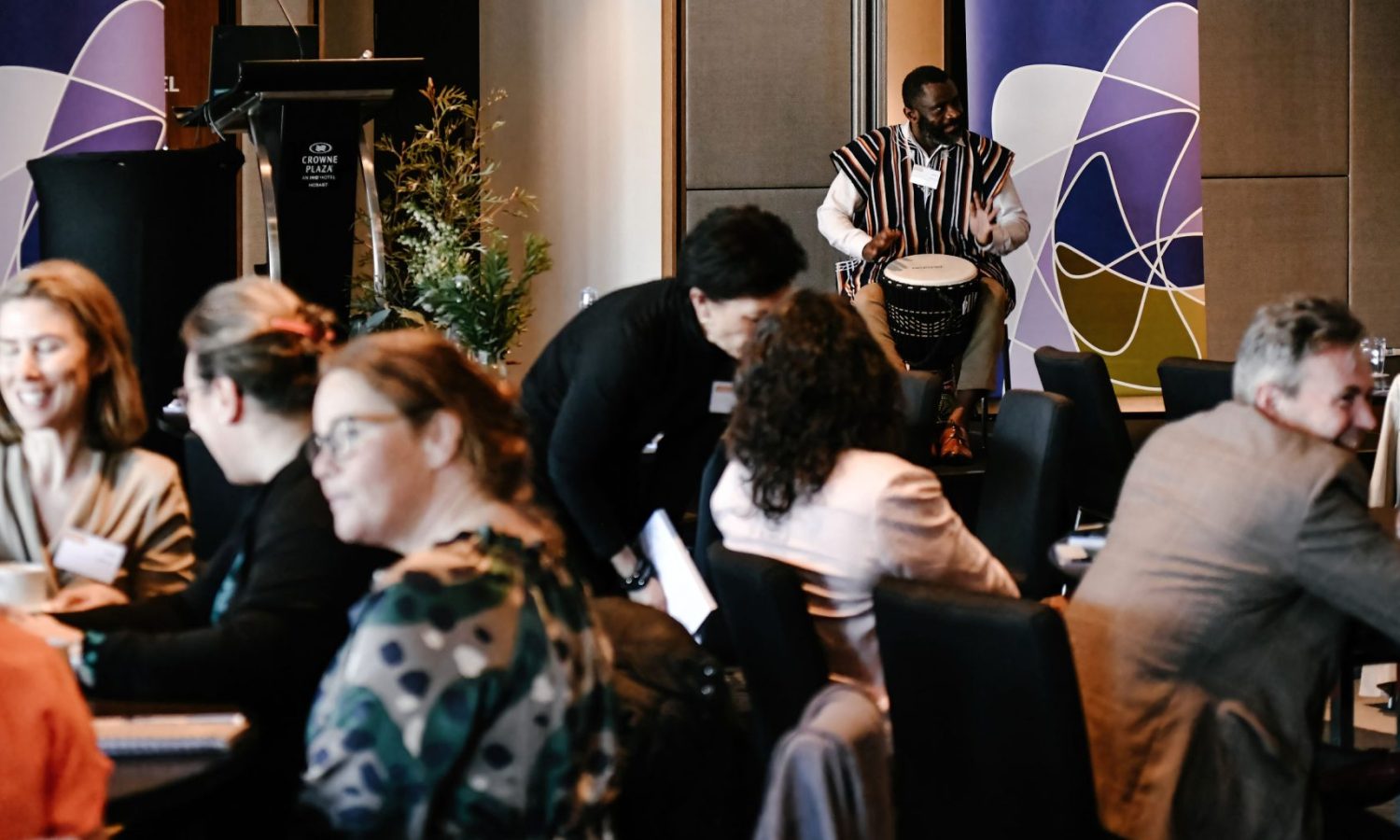 A landscape photo of a man playing hand drums on stage in front of members of the Symposium audience. There are Tasmanian Leaders banners and a lectern in the background.