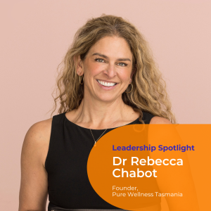 A portrait photo of Dr Rebecca Chabot. She stands against a light pink background and wears a sleeveless black dress with long curly blonde hair and smiles directly at camera.