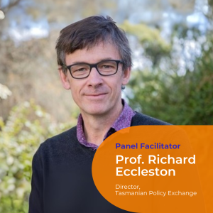 A portrait photo of Professor Richard Eccleston. He is outside with short dark hair, a purple shirt and blue jumper and black framed glasses. Green foliage background.