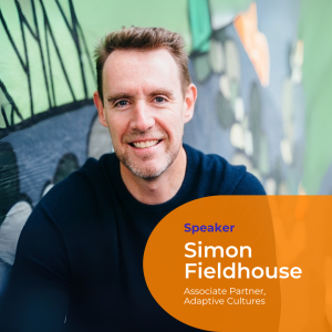 A portrait photo of Simon Fieldhouse. He sits in front of a green and blue wall. He has short spikey hair and wears a blue woollen jumper. He smiles directly at the camera.