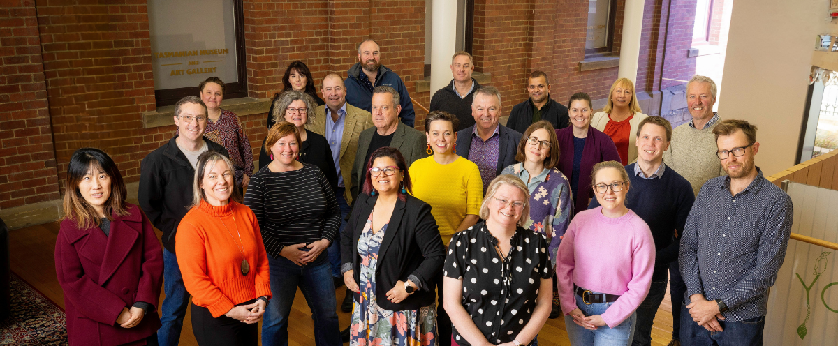 A landscape group photo of 23 of the 2024 Tasmanian Leaders program participants. All wearing brightly coloured professional wear, all smiling and looking directly at the camera. Brick wall background.