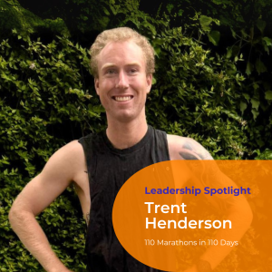 A portrait photo of Trent Henderson in a running singlet standing against a green hedge. He has blonde curly hair, tattoos down his left arm and smiles at the camera.