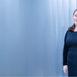 Picture of a woman standing against a Light blue background with her hair tied back wearing a dark grey dress with long black sleeves. her hand is in her pocket and she smiles at the camera.