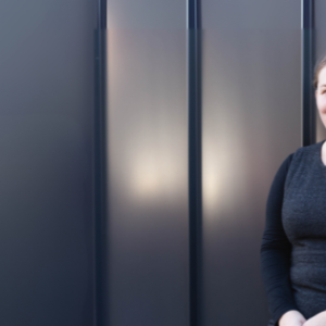 Picture of a woman standing against a dark grey steel wall with her hair tied back wearing a dark grey dress with long black sleeves. She is smiling at the camera.