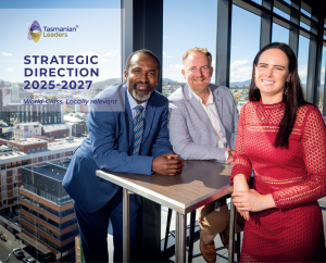 Photo of three TLP participants from 2023. A woman with long black hair and a long sleeved red dress, a fair haired man with a beard in a grey suit jacket and a dark haired man in a blue suit leaning on a small table together. View behind them overlooks the city of Hobart.