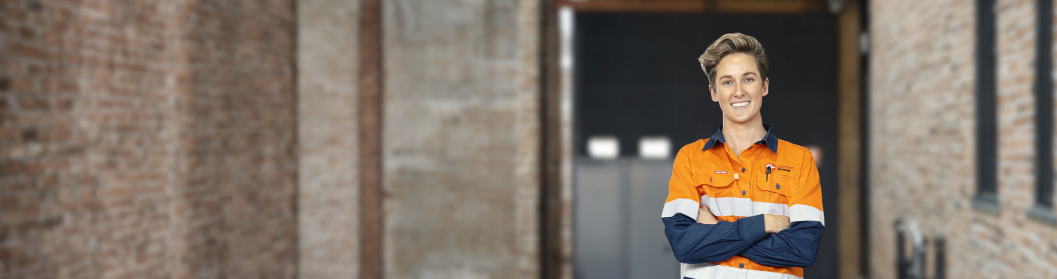 Image of a woman with short stylish hair, wearing a hi-vis workshirt. She stands with her arms crossed in front of an industrial site