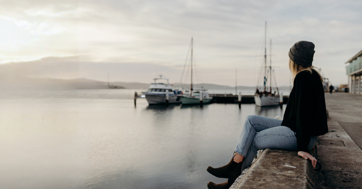 a woman is sitting on the edge of a wharf, looking away from the camera. In the distance there is a couple of boats moored at the wharf, and a mountain scape in the far background