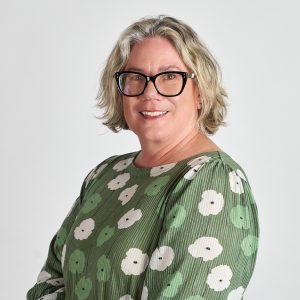 Ros Young headshot - a women with light bobbed wavy hair, glasses and a green printed top smiles at the camera