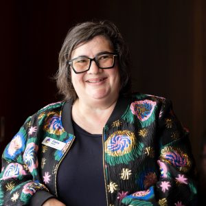 candid photo of Claire Beale - a woman with dark greying bobbed wavy hair and glasses, wearing a brocade bomber jacket and black top smiles at someone out of shot