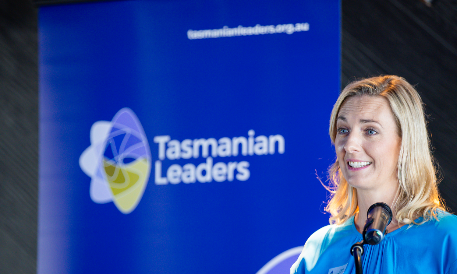 Woman speaking at a lectern at a Tasmanian Leaders event, with a large Tasmanian Leaders banner behind her.