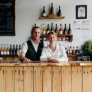 Two people smiling behind the timber cellar door counter at Eastford Creek Vineyard, surrounded by bottles of Tasmanian wine.