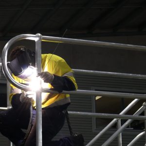 Worker in high-visibility clothing and protective welding equipment fabricating a large metal structure in an industrial workshop.